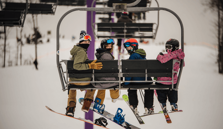 2 skiers and 2 snowboarders on the purple chairlift looking back at camera