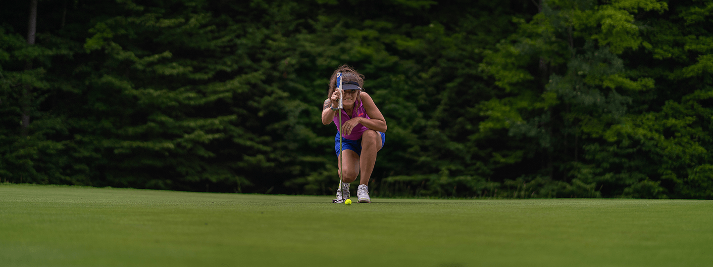 Woman golferr kneeling and sizing up a putt on the green of Hawk's Eye GC.