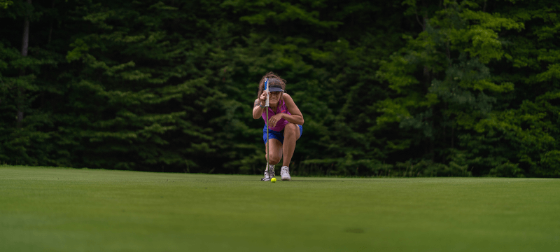 Woman golferr kneeling and sizing up a putt on the green of Hawk's Eye GC.