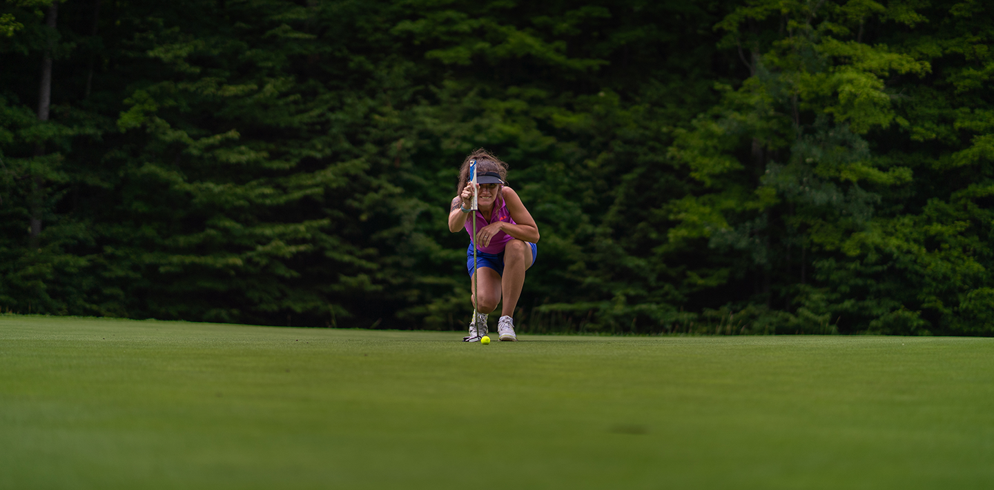 Woman golferr kneeling and sizing up a putt on the green of Hawk's Eye GC.