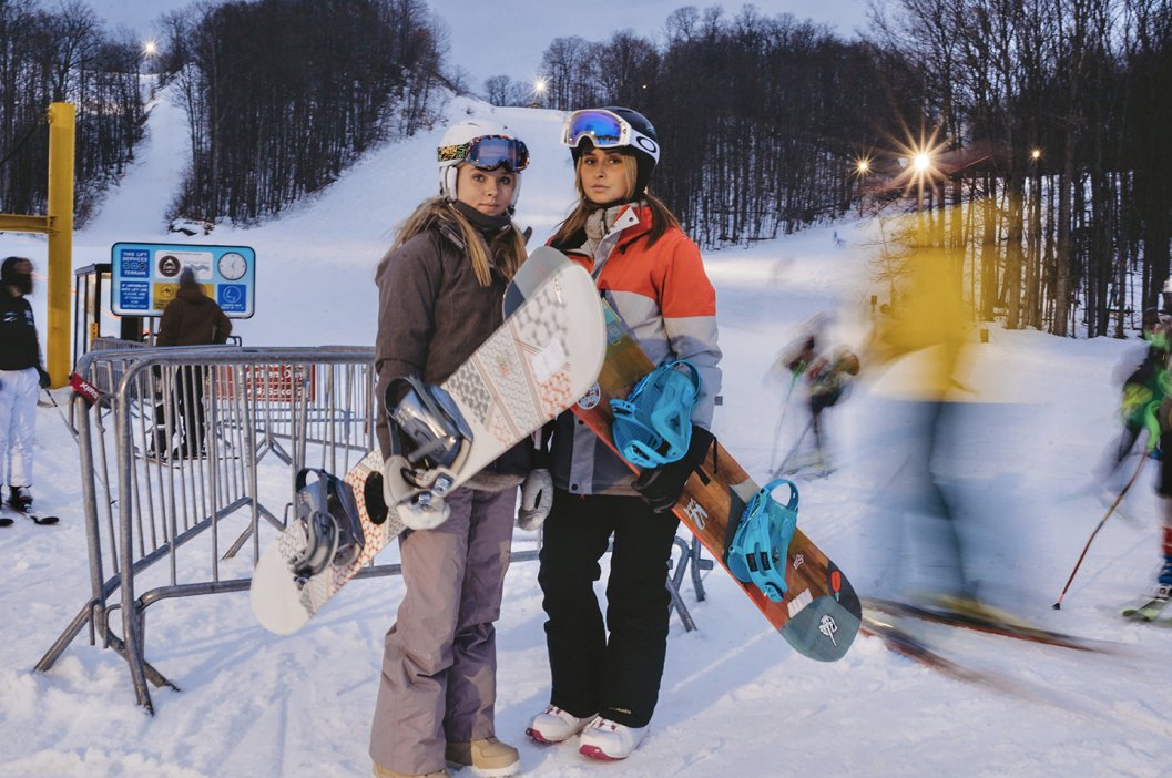 Two college aged women standing with their snowboards in hand in front of the yellow chairlift