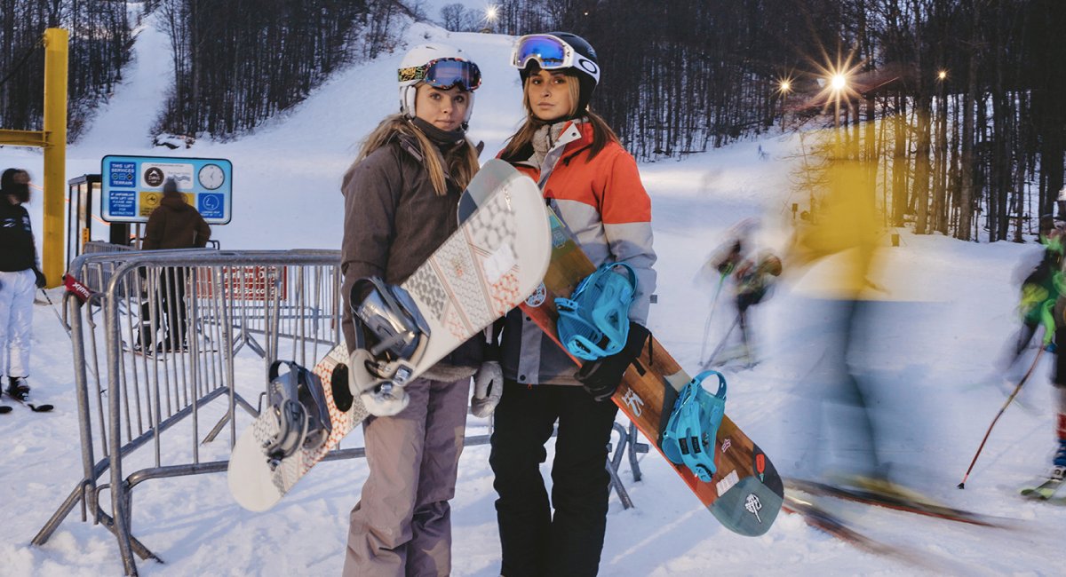 Two college aged women standing with their snowboards in hand in front of the yellow chairlift