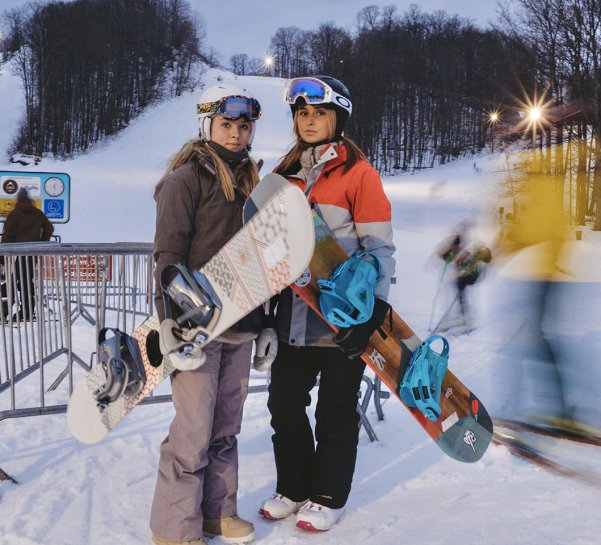 Two college aged women standing with their snowboards in hand in front of the yellow chairlift