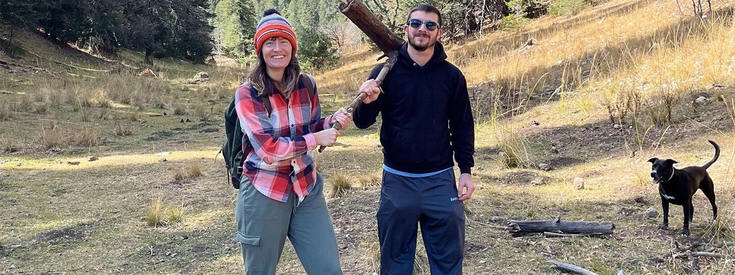 A happy couple hiking with their dog, holding a stick.